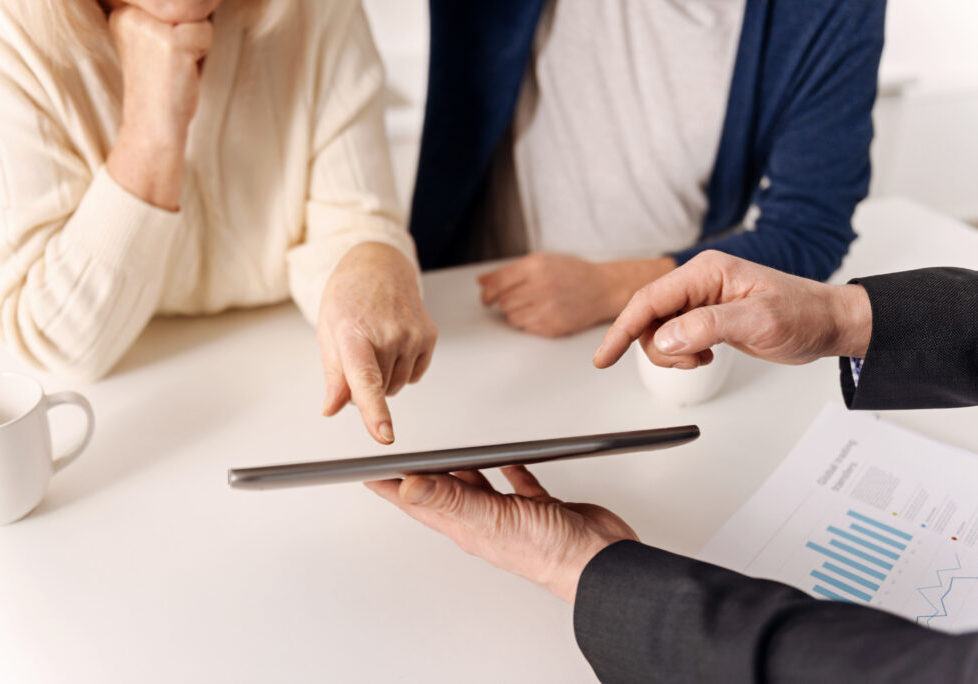Useful modern gadgets in our hands. Experienced mature skilled real estate agent working with aged couple of clients while expressing care and using tablet
